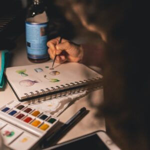 Artist painting with watercolors in a notepad at a desk. Creative process indoors.