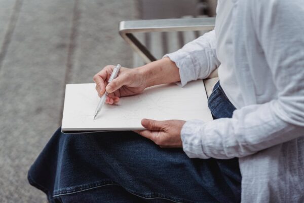 An adult drawing in a sketchbook while sitting outdoors, highlighting creativity and relaxation.