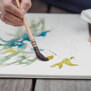 Artist's hands painting vibrant watercolor art in a sketchbook, capturing creativity and detail.