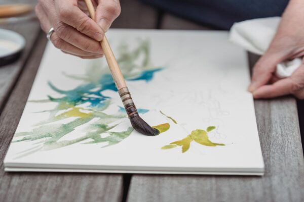 Artist's hands painting vibrant watercolor art in a sketchbook, capturing creativity and detail.